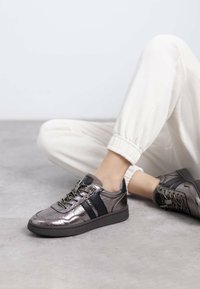 Zapatilla metálica gris con franjas negras; material texturizado, puntera redonda y suela plana. Cuenta con diseño de cordones y costuras de contraste claras.