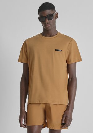 Joven con camiseta amarillo mostaza y pantalones cortos a juego, gafas de sol negras, de pie frente a un fondo liso gris claro.