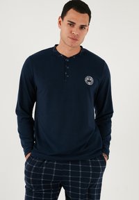 Cămașă henley cu mânecă lungă de culoare albastră închisă, cu gât butonat, având un material texturat și un logo circular pe piept, asortată cu pantaloni cu carouri.
