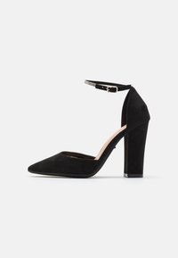 Chaussure à talons hauts en daim noir avec un bout pointu, un talon épais et une fine bride de cheville avec une petite boucle et un détail en rhinestones.