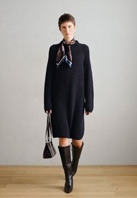 Πλεκτό μαύρο φόρεμα με μακριά μανίκια, υφασμάτινο σχέδιο, συνδυασμένο με ένα εμπριμέ κασκόλ, καφέ μπότες μέχρι το γόνατο και μια μικρή τσάντα.