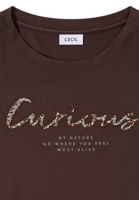 Chemise marron avec le mot « Curious » écrit en sequins argentés et dorés, et un texte en dessous disant « Par nature, va là où tu te sens le plus vivant. »