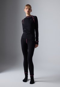 Woman standing in black fitted long-sleeve top and leggings with pink seam details on a gray gradient background.