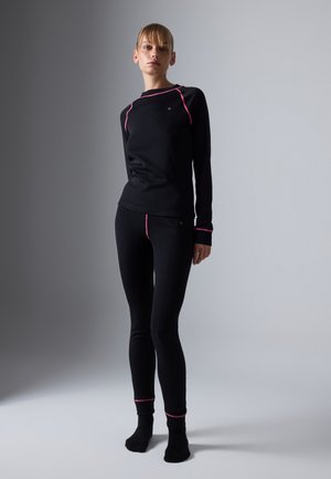 Woman standing in black fitted long-sleeve top and leggings with pink seam details on a gray gradient background.