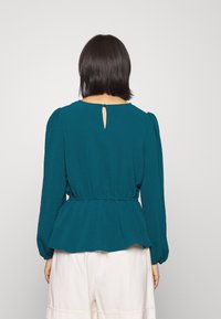 Donna con capelli corti e scuri che indossa una blusa a maniche lunghe color teal con cintura in vita e chiusura a bottone a forma di chiave, abbinata a pantaloni bianchi, girata di spalle.