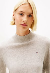 Jasno beżowy sweter dzianinowy z wysokim kołnierzem i prążkowanymi akcentami. Posiada małe logo w kolorach czerwonym, białym i niebieskim na klatce piersiowej.