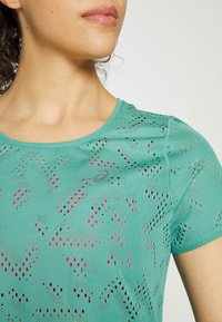 Turkusowa koszulka sportowa z dekoltem w serek, z oddychającymi wzorami wyciętymi laserowo w kolorze czerwonym i czarnym. Miękka tkanina o dopasowanym kroju.