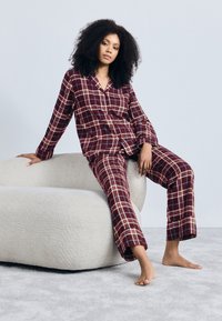 Σετ πιτζάμας σε κόκκινο και μπεζ καρό από μαλακό ύφασμα, με μπλούζα με κουμπιά και παντελόνι ίσιας γραμμής, φορεμένο ενώ κάθεται σε έναν καναπέ ανοιχτού χρώματος.