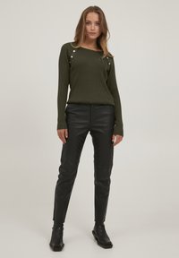 Pulover ribbed de culoare verde măsliniu cu accente din butoane argintii pe umeri, asortat cu pantaloni din piele negri și bocanci negri până la glezne.