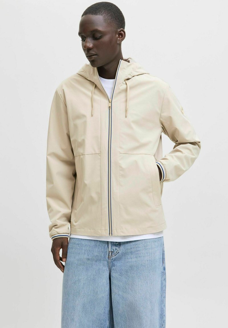 Beige, kapuzenjacke mit Reißverschluss, verfügt über zwei Fronttaschen und gestreifte Akzente an den Ärmelbündchen. Hergestellt aus glattem, wasserabweisendem Material.