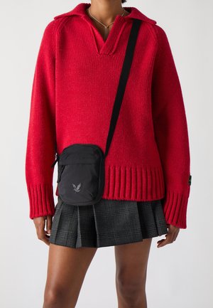 Pull en tricot rouge avec un col à revers, ourlet côtelé, associé à un sac bandoulière noir avec une poche zippée et un logo. Jupe grise à carreaux.