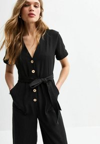 Combinaison noire à manches courtes, décolleté en V, fermeture à boutons sur le devant avec des boutons en bois, ceinture à la taille et poches sur les côtés. Texture du tissu lisse.