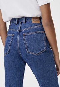 Blå denim mom jeans med hög midja, med en läderetikett markerad "Mom Jeans" på baksidan, och två bakfickor med synliga sömmar.