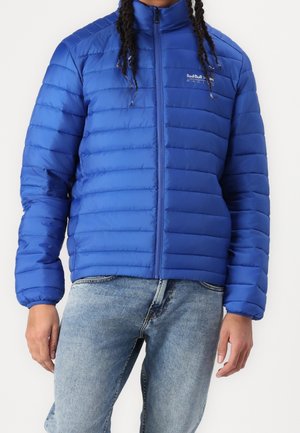 Person trägt eine blaue gesteppte Red Bull Racing Jacke kombiniert mit hellblauen Denim-Jeans, steht vor einem schlichten Hintergrund.