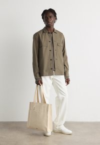 Olive green button-up shirt with two chest pockets, cream jeans, beige tote bag, and white sneakers, model standing against a neutral background.