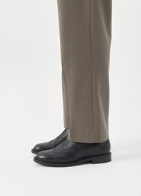 Sapatos de couro pretos com detalhes perfurados, combinados com calças cinzentas ajustadas. Os sapatos têm uma ponta arredondada e um salto baixo.