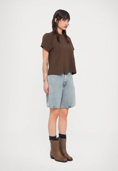 Κοντομάνικο καφέ τοπ, ανοιχτό μπλε τζιν σορτς και καφέ μποτάκια μέχρι τον αστράγαλο με μαύρες κάλτσες, εμφανίζονται μπροστά σε απλό λευκό φόντο.