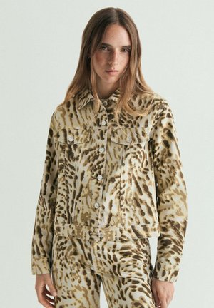 Joven con cabello largo que lleva una chaqueta y pantalones a juego con estampado animal en tonos beige y marrón, de pie contra un fondo claro y liso.