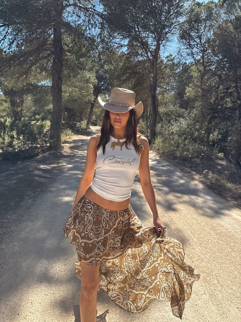 Brun paisleymønstret skjørt med volangkant, kombinert med en hvit ermeløs topp. Beige cowboyhatt gir et vestlig preg. Naturlig bakgrunn.