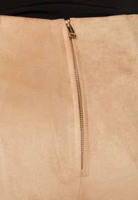 Beige semsket stoff med en jevn tekstur, med en synlig gullglidelås langs sidesømmen. Livbåndet er svart og elastisk.