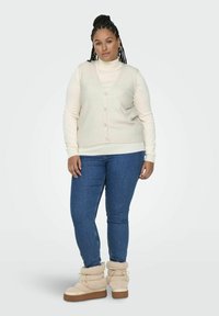 En gräddfärgad stickad väst över en vit polotröja, tillsammans med blå jeans och gräddfärgade stövlar med pälsfoder. Inkluderar knappar och ett strukturerat mönster.