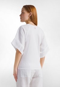 Mujer con cabello rojo que lleva una blusa blanca de manga corta y holgada y pantalones blancos, de pie de lado contra un fondo blanco liso.