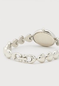 Ασημένιο μεταλλικό ρολόι χειρός με βραχιόλι από κρίκους σε σχήμα καρδιάς και κούμπωμα με το λογότυπο της Coach σε λευκό φόντο.