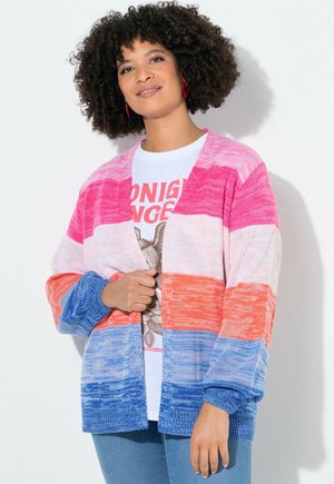 Žena s kudrnatými vlasy nosící červené náušnice, vícebarevný růžovo-oranžovo-modrý pruhovaný svetr a bílé tričko s potiskem.