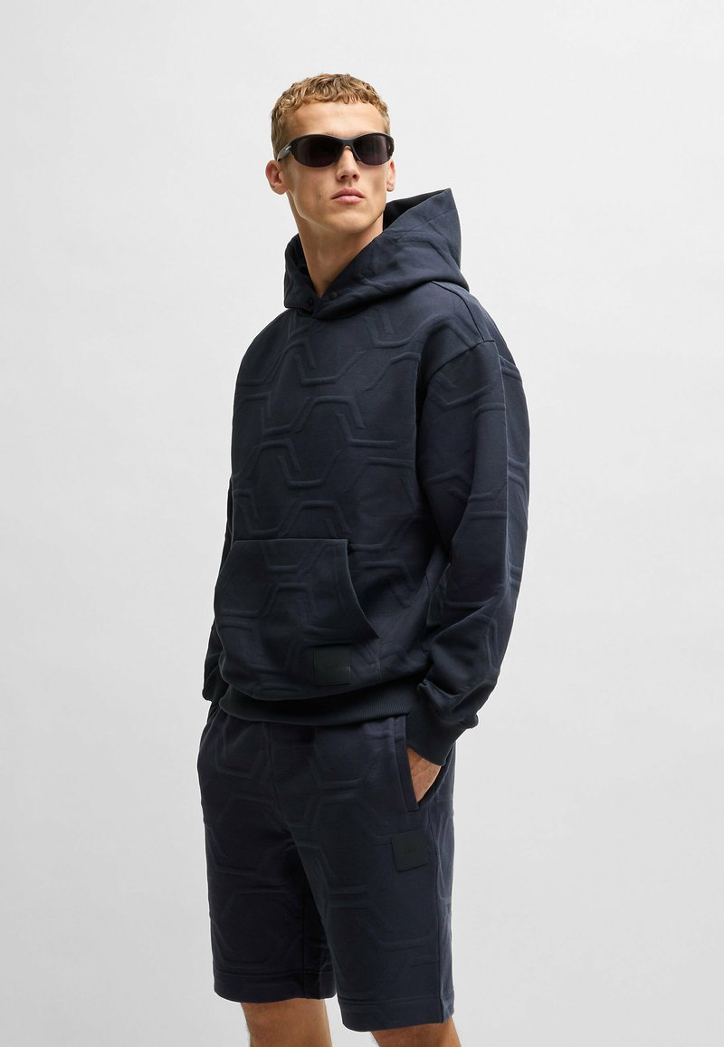 Navy blue hooded sweatshirt with a textured hexagonal pattern, paired with matching shorts, featured pockets and a subtle logo patch.