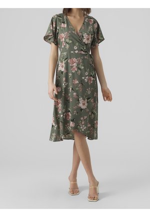 Femme portant une robe portefeuille verte à fleurs avec des manches volantées et des sandales à talons beige, debout devant un fond gris uni.