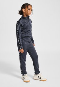 Granatowy dres z kołnierzem na suwak do ćwiartki, z białą taśmą z logo na rękawach i dopasowanymi, zwężanymi spodniami. Lekka i gładka tkanina.