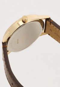 Montre en or avec un boîtier en acier inoxydable, un bracelet en cuir marron et un logo de marque gravé au dos. Comprend une couronne texturée.