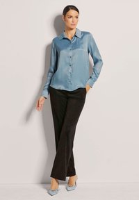 Lichtblauwe satijnen blouse met een kraag, knopen aan de voorkant, gecombineerd met zwarte broek en blauw met een strik versierde hakken. Soepele textuur.