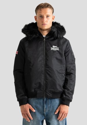 Jeune homme portant une veste noire Lonsdale avec capuche en fourrure, écusson Union Jack sur la manche, debout sur un fond uni gris clair.