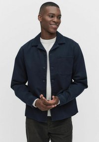 Mørkeblå knap-op jakke i robust stof med spids krave og enkelt brystlomme; parret med en lys sweater og mørke bukser.