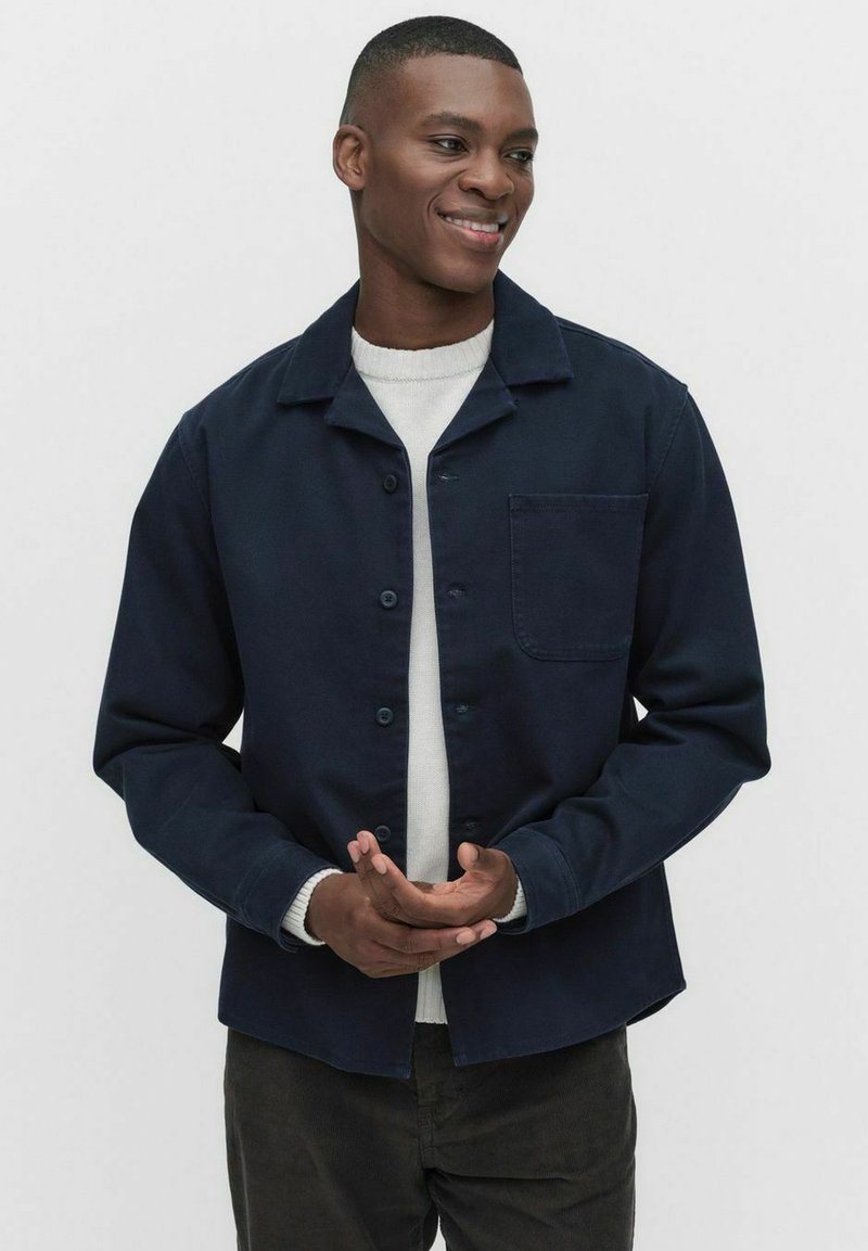 Donkerblauwe button-up jas van stevige stof met een puntige kraag en een enkele borstzak; gecombineerd met een lichte trui en donkere broek.