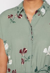 Grünes Blumenkleid mit kurzen Ärmeln, das mit roten und weißen Blumenmustern verziert ist, einen kragenförmigen Ausschnitt hat und mit Knöpfen geschlossen wird. Weicher, strukturierter Stoff.