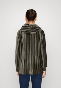 Giacca di velluto verde con pieghe verticali, cappuccio e polsini a coste, indossata con jeans blu; presenta una texture morbida e una leggera lucentezza.