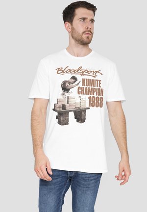 Man wearing white t-shirt with graphic of martial artist breaking bricks and text "Bloodsport Kumite Champion 1988" standing against gray background.