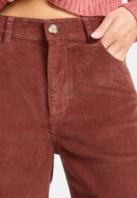 Pantaloni in corda color ruggine con righe verticali, dotati di chiusura frontale con bottone e tasche laterali. La trama del tessuto offre una finitura morbida.