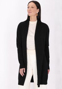 Čierny cardigan s dlhými rukávmi, otvoreným predným dielom, rebrovanými manžetami a jemnou textúrou. Nosený cez svetlý top a krémové nohavice.