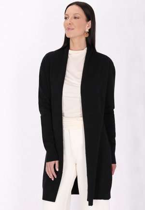 Cardigan nero con maniche lunghe, apertura anteriore, polsini a coste e una sottile texture. Indossato sopra un top chiaro e pantaloni color crema.