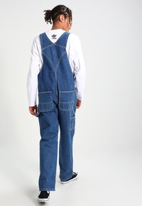 Demi overall berwarna biru, dilengkapi dengan tali yang dapat disesuaikan, dua saku depan, dan potongan longgar. Dipadukan dengan kemeja lengan panjang putih dan sepatu hitam.