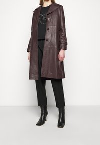 Trench in pelle color bordeaux con ampio collo, doppiopetto con bottoni e tasche laterali, abbinato a una maglia nera e pantaloni su misura. Stivali neri con tacco.
