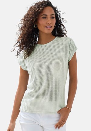 Woman with curly hair wearing gold hoop earrings, a light textured short-sleeve top, white pants, and a thin bracelet, posing with hand in pocket.