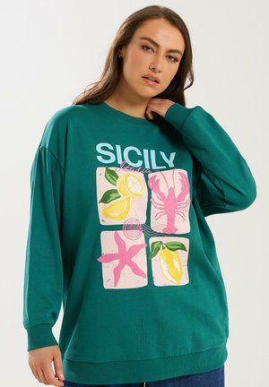 Vrouw draagt een teal sweatshirt met een kleurrijk Siciliaans ontwerp met citroenen, een kreeft en een zeester, staand tegen een effen achtergrond.