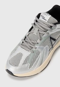 Zapatillas deportivas con parte superior de malla en plata y gris, con cordones grises, suela texturizada y detalles en negro. Diseño ligero para mayor comodidad.