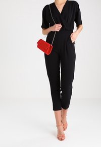 Czarny kombinezon z fasonem kopertowym i krótkimi rękawami, wyposażony w kieszenie z przodu. Połączony z czerwoną torebką z brokatem i cielistymi sandałami na paski.