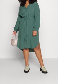 Zelené midi šaty s véčkovým výstřihem, zapínáním na knoflíky vpředu, dlouhými rukávy a stahováním v pase. Obsahují kapsu a postranní rozparky. V kombinaci se světlými botami.