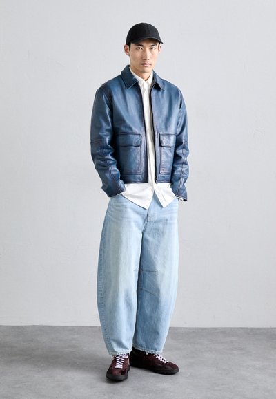 Un uomo è in piedi contro una parete semplice indossando un berretto nero, una giacca di pelle blu, una camicia bianca, jeans larghi di colore azzurro chiaro e scarpe scure con lacci bianchi.